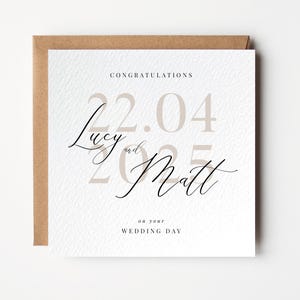 Puede incluir: Una tarjeta de boda blanca con ribete marrón. La tarjeta tiene el texto "Felicidades" en la parte superior, seguido de la fecha "22.04.2025" en números grandes de color beige. Los nombres "Lucy y Matt" están escritos en cursiva debajo de la fecha. El texto "En su día de boda" está en la parte inferior de la tarjeta.