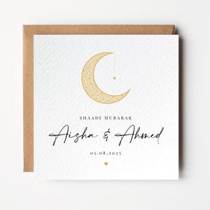 Pu&ograve; includere: Biglietto di nozze bianco con un design a mezzaluna e stella dorate. La carta include il testo "SHAADI MUBARAK" sopra i nomi "Aisha & Ahmed" e la data "05.08.2025". Un piccolo cuore dorato in basso.