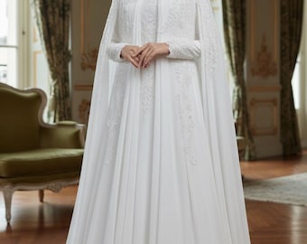 May include: A white wedding dress with a long, flowing skirt and a matching cape. The dress features intricate lace detailing on the bodice and sleeves. The model is wearing a white hijab. The dress is suitable for a formal event.