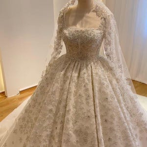 以下が含まれることがあります： 長いトレーンとベールが付いた白いウェディングドレス。ドレスはスパンコールとビーズで覆われており、キラキラと輝いています。ドレスはスクエアネックラインとロングスリーブです。