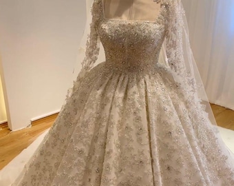 Puede incluir: Un vestido de novia blanco con una cola larga y un velo. El vestido está cubierto de lentejuelas y cuentas, creando un efecto brillante. El vestido tiene un escote cuadrado y mangas largas.