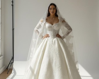 Pode incluir: Um vestido de noiva branco com decote em coração e mangas compridas, adornado com rendas delicadas. O vestido tem uma saia ampla e é complementado por um véu a condizer. A modelo posa num estúdio.