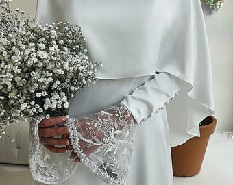 May include: A white satin dress with a cape and lace sleeves. The sleeves are trimmed with pearls. The dress is being held by a bouquet of white flowers.
