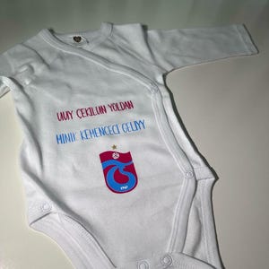 May include: White baby onesie with a blue and red logo and the text "UYUY ÇEKILIN YOLDAN MINIK KEMENCECI GELIY" in Turkish.