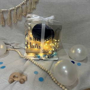 May include: A clear gift box tied with a white ribbon, containing a dark blue object and fairy lights. The box has text that reads "Omer Halis Sunnet oldu". Decorative elements include white balloons, wooden beads, and a small wooden car.