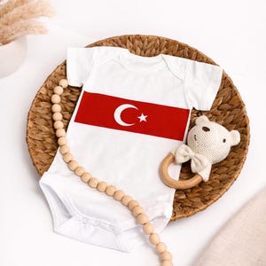 Könnte beinhalten: Weißer Baby-Body mit rotem Streifen, der einen Mond und einen Stern zeigt. Der Body liegt auf einem geflochtenen Korb, zusammen mit einer Holzperlen-Girlande und einer gestrickten Bärenrassel.