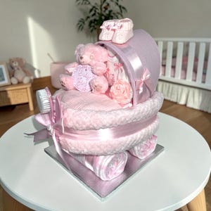 May include: A baby gift set shaped like a pink pram, constructed from rolled fabric items. It features a plush pink teddy bear holding a round tag that reads Baby Girl, a pair of small knitted booties, a hairbrush, and a decorative fabric flower.
