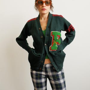 May include: Dark green vintage cardigan with a large letter "F" patch in red and green. The cardigan has red stripes on the sleeves and a button-up front. The person is wearing plaid pants and sunglasses.
