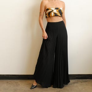 May include: A strapless tube top featuring a swirling abstract pattern in shades of brown, black, and white, paired with high-waisted black wide-leg trousers that feature vertical pleats throughout the fabric, extending down to the floor.