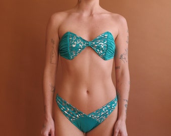 Puede incluir: Un top y una parte inferior de bikini verde azulado con estampado de leopardo plateado. La parte superior es de estilo bandeau con un lazo en la parte delantera. La parte inferior es de estilo de corte alto con un frente en forma de V.