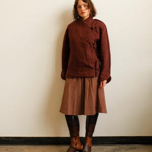 May include: A dark maroon textured jacket with a side-fastening toggle closure, paired with a solid brown knee-length skirt and two-toned brown leather knee-high boots featuring a dark brown upper section and a lighter brown lower foot area.