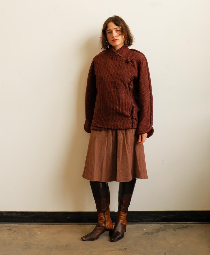 May include: A dark maroon textured jacket with a side-fastening toggle closure, paired with a solid brown knee-length skirt and two-toned brown leather knee-high boots featuring a dark brown upper section and a lighter brown lower foot area.