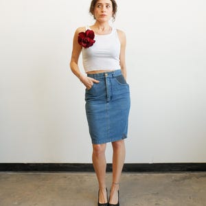 May include: A woman wearing a white tank top with a large red flower pinned to the shoulder, paired with a denim pencil skirt and black heels. The skirt is a medium wash denim. The background is a plain white wall.