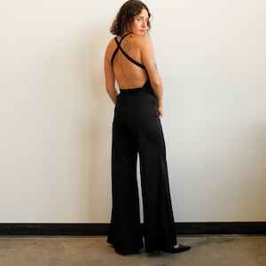 May include: A solid black jumpsuit featuring wide-leg trousers and a sleeveless bodice with a deep open back. The garment includes thin black straps that cross over the back, creating a distinct X-shape design above the waistline.