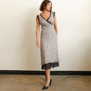 May include: A sleeveless midi dress featuring a cream base with a small black floral print, a V-neckline with black lace trim, and a layered black sheer hem. The dress has a fitted bodice and a straight skirt silhouette that falls below the knees.
