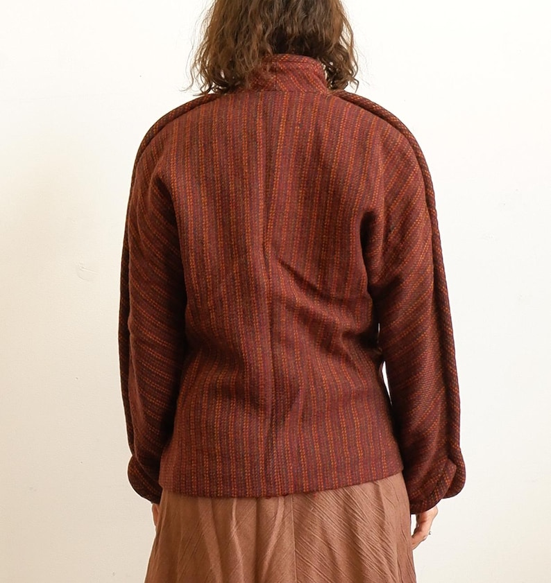 May include: A long-sleeved jacket in a deep rust and dark brown vertical striped pattern. The jacket features a high collar, structured shoulders, and a straight hemline, paired with a solid light brown skirt with a subtle textured finish.