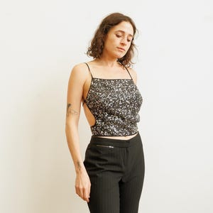 Op de afbeelding: Een zwarte crop top met pailletten, dunne bandjes en een vierkante halslijn. De top heeft een zilver en zwart paillettenpatroon. Het model draagt een zwarte broek met krijtstreep. De top heeft uitgesneden zijkanten.