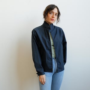 May include: A navy blue jacket with a high collar and a full-length zipper. The jacket has long sleeves and is worn over a light green top and blue jeans. The jacket is a solid color and appears to be made of a lightweight material.