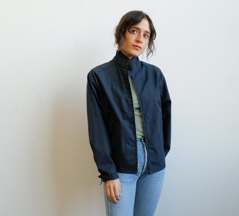 May include: A navy blue jacket with a high collar and a full-length zipper. The jacket has long sleeves and is worn over a light green top and blue jeans. The jacket is a solid color and appears to be made of a lightweight material.