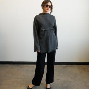 May include: A person wearing a dark gray, long-sleeved sweater with a high neckline and a pocket detail. The sweater has a frayed hem. The person is also wearing black wide-leg pants and black pointed-toe shoes.