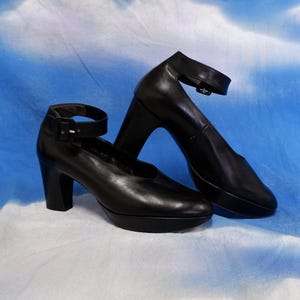 May include: A pair of black platform heels with ankle straps. The shoes have a rounded toe and a thick, block heel. The ankle straps fasten with a small buckle. The shoes are made of a smooth, shiny material.