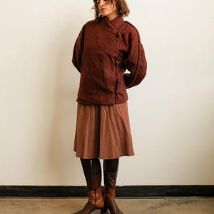 May include: A dark maroon textured jacket with a high collar and side toggle closures, paired with a solid brown knee-length skirt and two-toned brown leather boots featuring a square toe and a block heel, creating a layered ensemble.