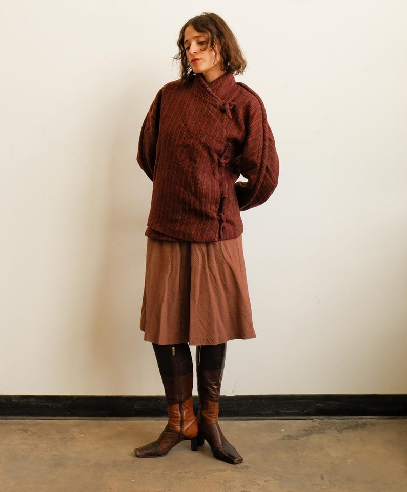 May include: A dark maroon textured jacket with a high collar and side toggle closures, paired with a solid brown knee-length skirt and two-toned brown leather boots featuring a square toe and a block heel, creating a layered ensemble.