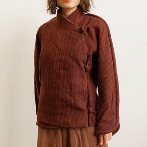 May include: A long-sleeved jacket in a deep burgundy and orange striped pattern. The jacket features a high collar, a wrap-style front closure secured with fabric ties, and a textured weave. It is paired with a solid brown skirt.