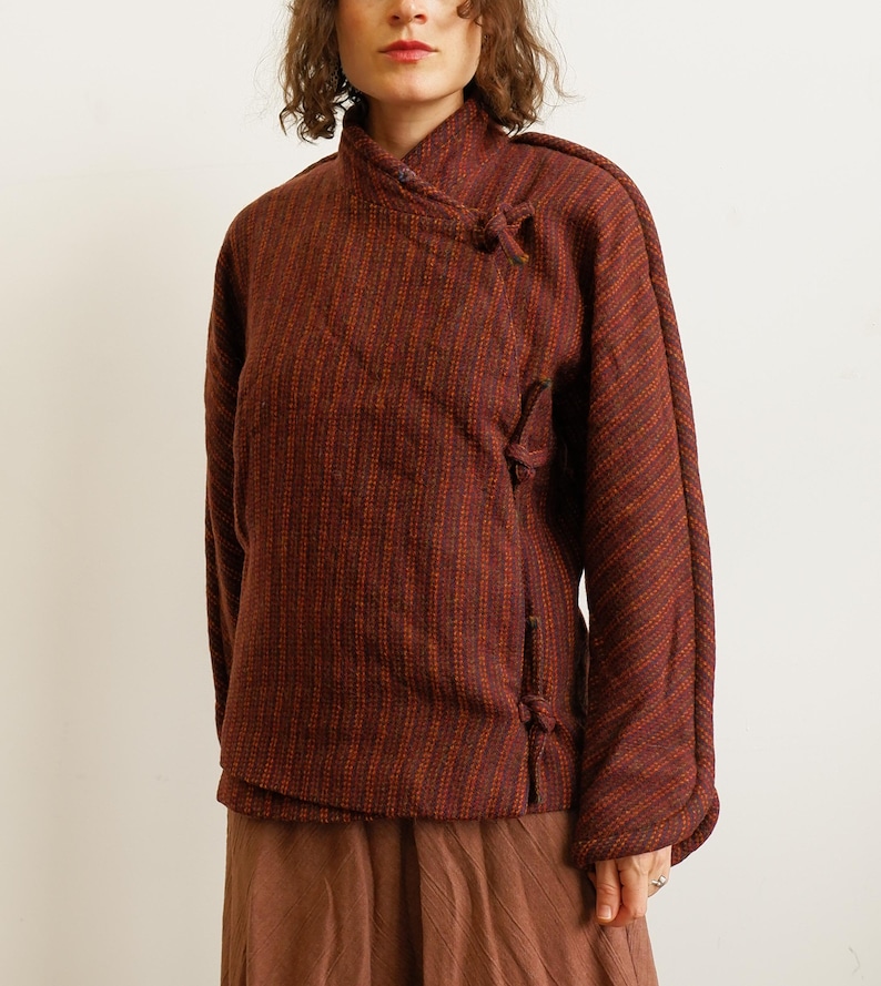 May include: A long-sleeved jacket in a deep burgundy and orange striped pattern. The jacket features a high collar, a wrap-style front closure secured with fabric ties, and a textured weave. It is paired with a solid brown skirt.