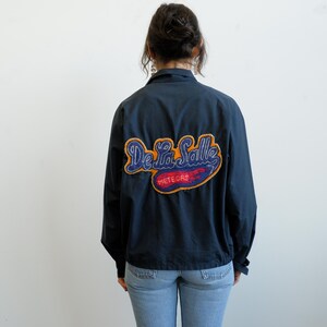 May include: Navy blue jacket with a colorful embroidered patch on the back. The patch features the words "De La Salle" in orange and blue script, with "METEORS" below in red and white. The jacket is worn with light blue jeans.
