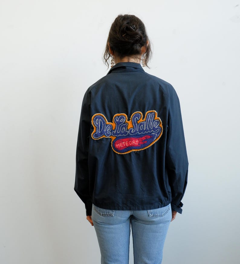 May include: Navy blue jacket with a colorful embroidered patch on the back. The patch features the words "De La Salle" in orange and blue script, with "METEORS" below in red and white. The jacket is worn with light blue jeans.