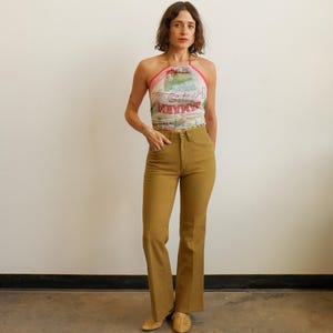 Può includere: Un top all'americana con una stampa grafica colorata con il testo Jamaica e Isle of Springs, abbinato a pantaloni color marrone chiaro a vita alta, taglio dritto e chiusura con bottone. L'outfit è completato da scarpe chiuse color marrone chiaro.