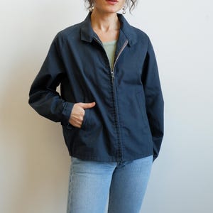 May include: A navy blue jacket with a full-length zipper, a collared neckline, and two front pockets. The jacket is made of a smooth fabric and has long sleeves. The person in the image is wearing light blue jeans.