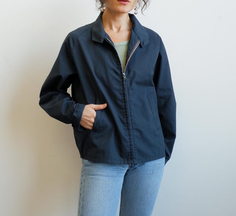 May include: A navy blue jacket with a full-length zipper, a collared neckline, and two front pockets. The jacket is made of a smooth fabric and has long sleeves. The person in the image is wearing light blue jeans.