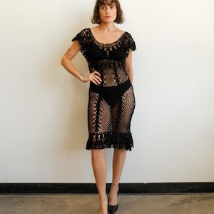 May include: A black knee-length dress crafted from intricate crochet with a wide scoop neckline, short cap sleeves, and a semi-sheer openwork mesh pattern throughout. The garment features a fitted bodice and a flared hemline with decorative trim.