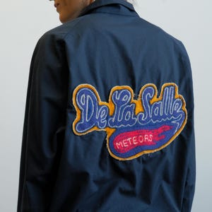 May include: Navy blue jacket with a large embroidered patch on the back. The patch features the words "De La Salle" in blue script, with "Meteors" in red, and a yellow border.