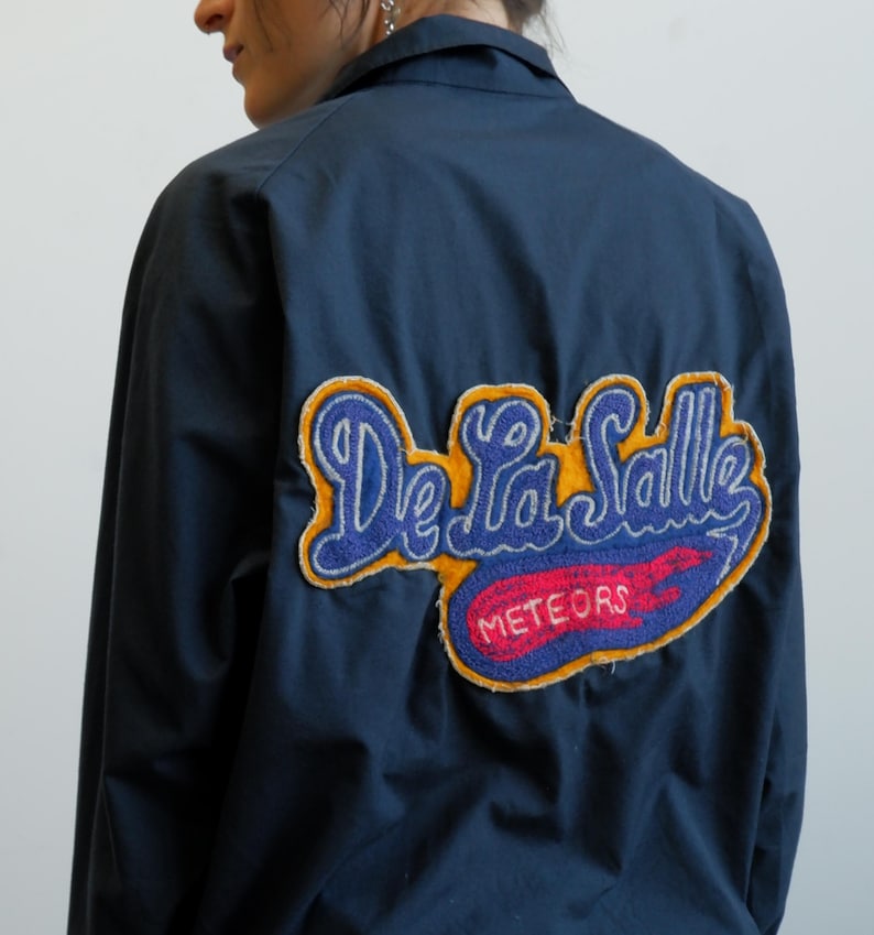 May include: Navy blue jacket with a large embroidered patch on the back. The patch features the words "De La Salle" in blue script, with "Meteors" in red, and a yellow border.