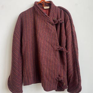 May include: A long-sleeved jacket in a deep burgundy and orange striped pattern. The garment features a high collar, a wrap-style front closure secured with three fabric knot toggles, and a solid burgundy interior lining visible at the neckline.