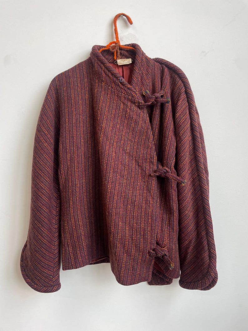 May include: A long-sleeved jacket in a deep burgundy and orange striped pattern. The garment features a high collar, a wrap-style front closure secured with three fabric knot toggles, and a solid burgundy interior lining visible at the neckline.