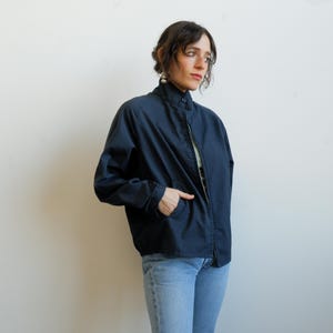 May include: A navy blue jacket with a high collar and a zipper closure. The jacket has two front pockets and is worn with light blue jeans. The jacket is a casual style.