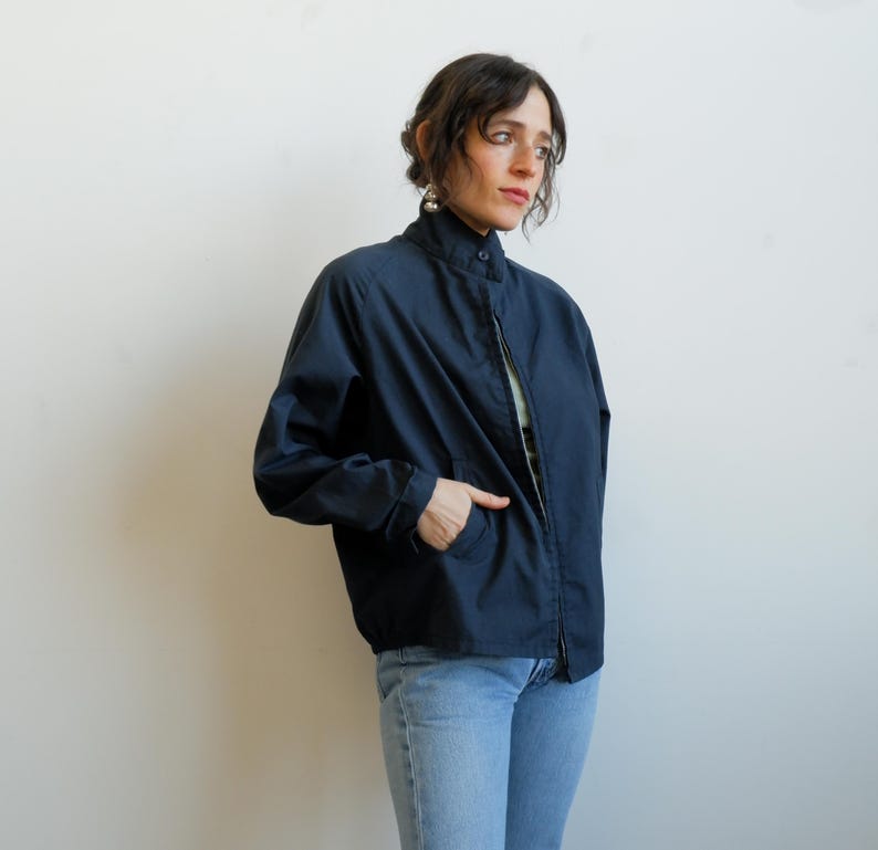 May include: A navy blue jacket with a high collar and a zipper closure. The jacket has two front pockets and is worn with light blue jeans. The jacket is a casual style.