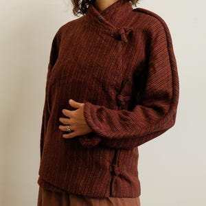 May include: A long-sleeved jacket in a deep rust and dark brown textured weave. The garment features a high collar and an asymmetrical front closure secured by three fabric-covered toggle knots. The fabric has a subtle vertical striped pattern.