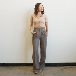 May include: A woman wearing a light brown short-sleeved cardigan with black trim and buttons, paired with high-waisted, wide-leg gray trousers. The outfit is completed with beige shoes. The background is a plain white wall.