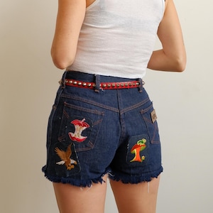 May include: Dark blue denim shorts with frayed edges, featuring embroidered patches of an apple core, an eagle, and a mushroom. A red studded belt is worn with the shorts. The shorts have a high waist and a classic five-pocket design.