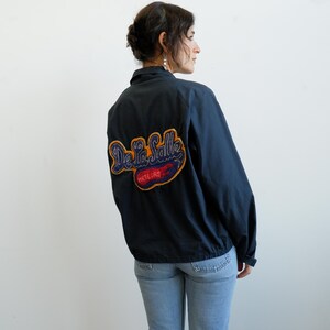 May include: Navy blue jacket with a large embroidered patch on the back that reads "De La Salle Meteors" in a script font. The patch has blue, red, and gold colors. The jacket has a collar and a zipper closure.