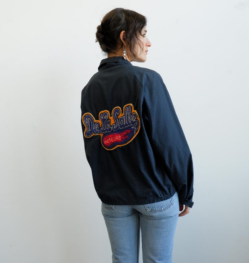 May include: Navy blue jacket with a large embroidered patch on the back that reads "De La Salle Meteors" in a script font. The patch has blue, red, and gold colors. The jacket has a collar and a zipper closure.