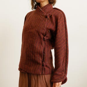 May include: A long-sleeved jacket in a deep rust and dark brown textured fabric with a subtle diagonal stripe pattern. The jacket features a high collar and a side-fastening closure secured by three fabric-covered toggle knots along the front.