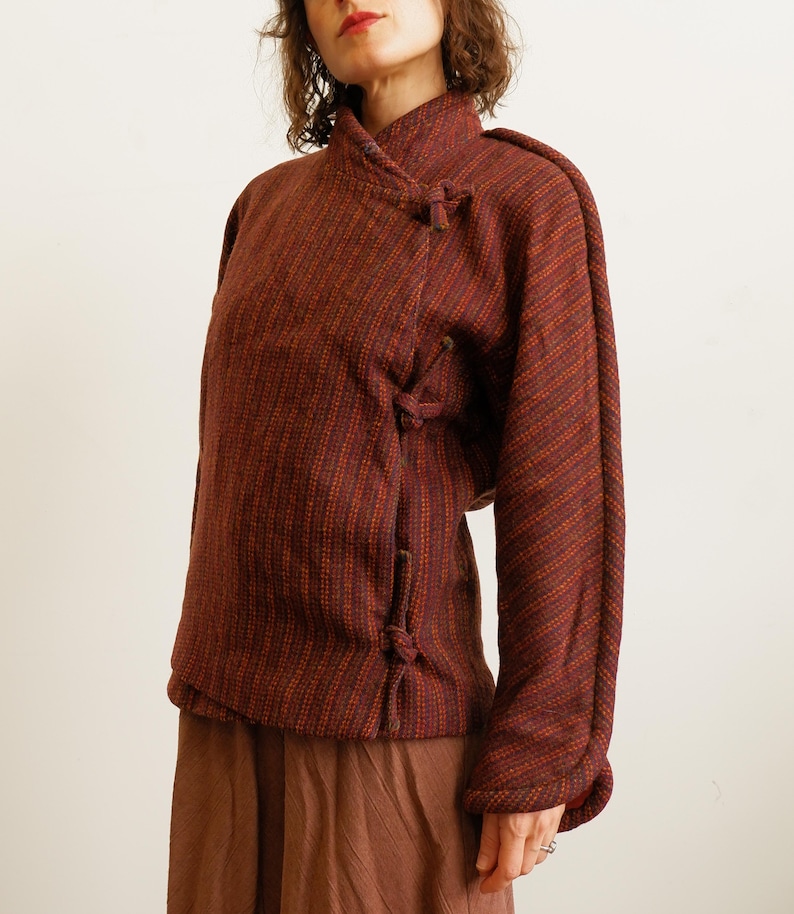 May include: A long-sleeved jacket in a deep rust and dark brown textured fabric with a subtle diagonal stripe pattern. The jacket features a high collar and a side-fastening closure secured by three fabric-covered toggle knots along the front.