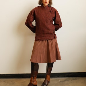May include: A dark red and black striped wrap jacket with a high collar and toggle closures, paired with a solid brown knee-length skirt and two-toned brown and black leather knee-high boots with a block heel and side zipper closure.