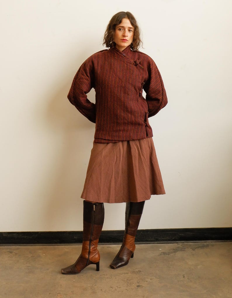 May include: A dark red and black striped wrap jacket with a high collar and toggle closures, paired with a solid brown knee-length skirt and two-toned brown and black leather knee-high boots with a block heel and side zipper closure.